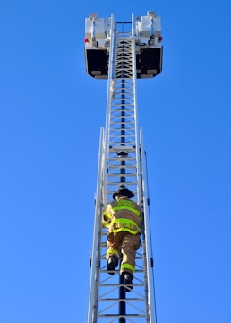 A firefighter is climbing a tall metal ladder on a fire truck, reaching towards two platforms against a clear blue sky. The firefighter is wearing protective gear, including a helmet and high-visibility jacket.