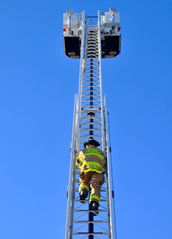 A firefighter is climbing a tall metal ladder on a fire truck, reaching towards two platforms against a clear blue sky. The firefighter is wearing protective gear, including a helmet and high-visibility jacket.