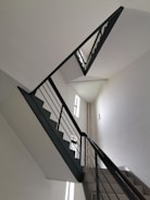 A staircase with a modern design, featuring clean, angular lines and dark railing against white walls. The stairs ascend in a zigzag pattern, and natural light filters in through narrow windows on the left.