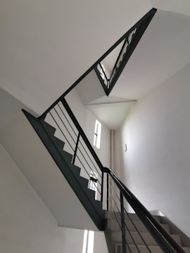 A staircase with a modern design, featuring clean, angular lines and dark railing against white walls. The stairs ascend in a zigzag pattern, and natural light filters in through narrow windows on the left.