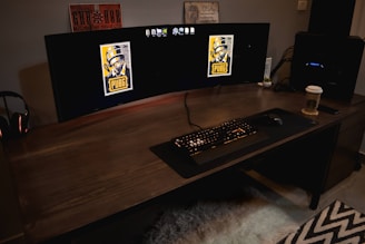 A gaming setup featuring a large curved monitor displaying PUBG logos on the screens. A keyboard and mouse are positioned on a large desk pad. A pair of headphones rests on the desk alongside a mobile phone. A drink in a Starbucks cup is placed to the right, while a desktop tower is seen in the background on the right side.