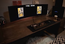 A gaming setup featuring a large curved monitor displaying PUBG logos on the screens. A keyboard and mouse are positioned on a large desk pad. A pair of headphones rests on the desk alongside a mobile phone. A drink in a Starbucks cup is placed to the right, while a desktop tower is seen in the background on the right side.
