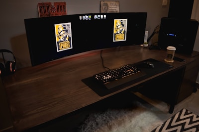 A gaming setup featuring a large curved monitor displaying PUBG logos on the screens. A keyboard and mouse are positioned on a large desk pad. A pair of headphones rests on the desk alongside a mobile phone. A drink in a Starbucks cup is placed to the right, while a desktop tower is seen in the background on the right side.