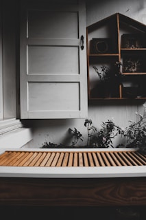 A rustic setting featuring a white wooden window with a simple handle. Below the window is a brown slatted wooden surface, possibly a bench or table. To the right, a wooden shelf with a house-shaped design holds small decorative items. Some green plants and foliage are visible around the shelf, adding a touch of nature.