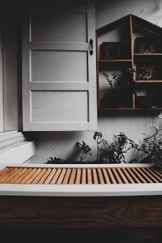 A rustic setting featuring a white wooden window with a simple handle. Below the window is a brown slatted wooden surface, possibly a bench or table. To the right, a wooden shelf with a house-shaped design holds small decorative items. Some green plants and foliage are visible around the shelf, adding a touch of nature.