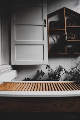 A rustic setting featuring a white wooden window with a simple handle. Below the window is a brown slatted wooden surface, possibly a bench or table. To the right, a wooden shelf with a house-shaped design holds small decorative items. Some green plants and foliage are visible around the shelf, adding a touch of nature.