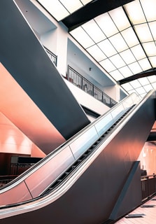 An indoor space featuring an escalator in a modern architectural setting. The escalator is angled upward, and there are geometric, grid-like patterns on the ceiling. Soft lighting enhances the sleek design of the area, and the color palette includes a warm pink hue contrasting with darker shades.