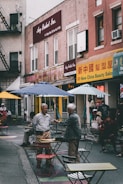 A bustling urban street scene features people interacting among scattered tables and chairs outside. Two men are engaged in conversation in the foreground, while others sit or walk by. The background displays signs with a mix of English and Chinese characters, suggesting a multicultural neighborhood setting.