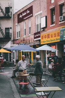 A bustling urban street scene features people interacting among scattered tables and chairs outside. Two men are engaged in conversation in the foreground, while others sit or walk by. The background displays signs with a mix of English and Chinese characters, suggesting a multicultural neighborhood setting.