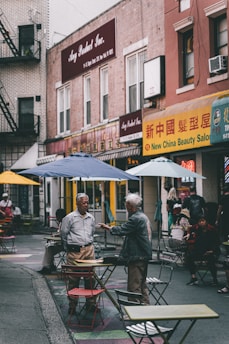 A bustling urban street scene features people interacting among scattered tables and chairs outside. Two men are engaged in conversation in the foreground, while others sit or walk by. The background displays signs with a mix of English and Chinese characters, suggesting a multicultural neighborhood setting.
