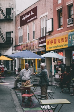 A bustling urban street scene features people interacting among scattered tables and chairs outside. Two men are engaged in conversation in the foreground, while others sit or walk by. The background displays signs with a mix of English and Chinese characters, suggesting a multicultural neighborhood setting.