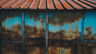 A weathered and rusted metal sheet is supported by vertical wooden beams, featuring a combination of blue paint and brown rust stains. The metal is topped with a corrugated roof of reddish-brown tin.