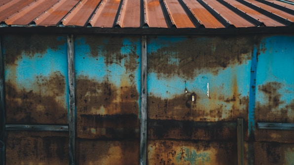 A weathered and rusted metal sheet is supported by vertical wooden beams, featuring a combination of blue paint and brown rust stains. The metal is topped with a corrugated roof of reddish-brown tin.