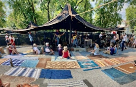 A group of people are gathered outdoors under a large black canopy to watch musicians playing on stage. The setting is casual, with attendees sitting on colorful rugs spread over gravel. Trees in the background provide a natural atmosphere, and people are dressed comfortably, indicating a relaxed, warm weather event.