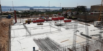 A construction site shows an early stage of building, featuring numerous steel reinforcements and foundations. Workers are visible in the background, with industrial equipment such as a digger and various materials including pipes and boards scattered around. In the background, the sea and hills can be seen, indicating a coastal location.