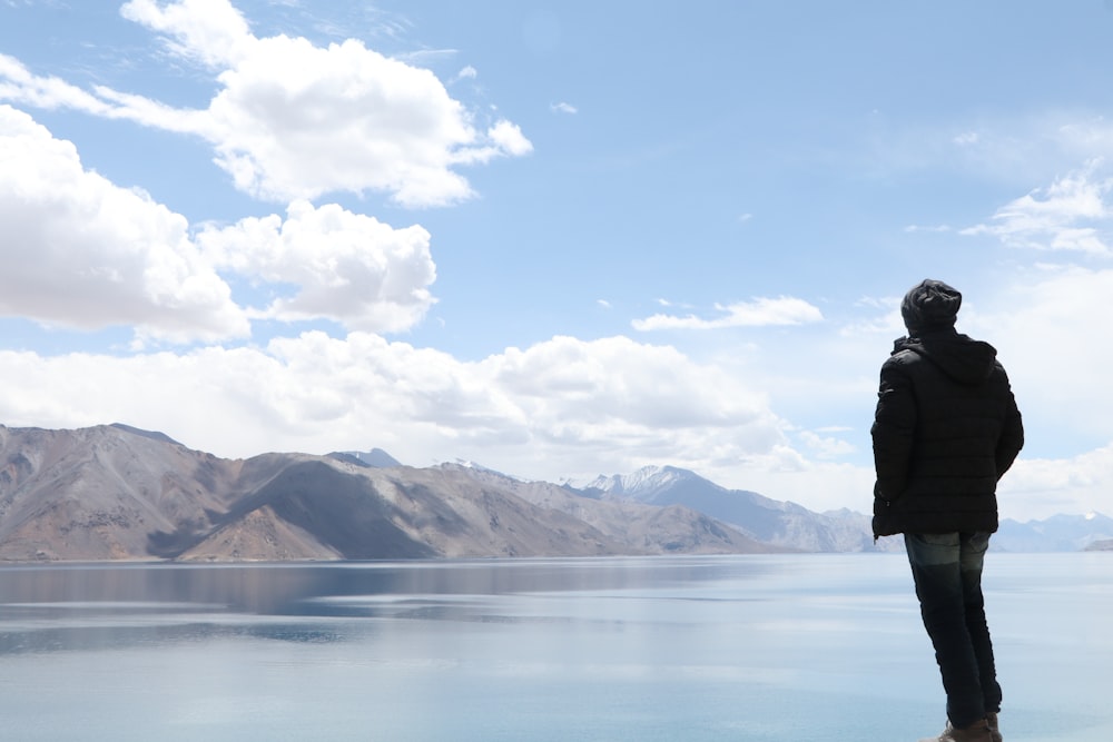 man standing beside water looking towards mountain