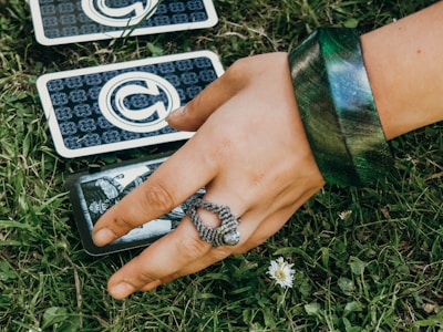 A hand adorned with a large green bracelet and a detailed silver ring explores tarot cards laid on lush green grass. Daisies are interspersed in the surroundings, adding a natural element. Tarot card designs are visible, indicating a mystical or fortune-telling theme.