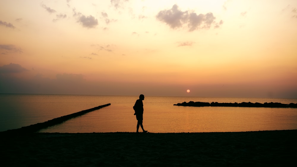 Silhouettenfotografie eines Mannes, der während der goldenen Stunde an der Küste entlang geht