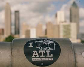 A sticker with a camera graphic and the text 'I ATL' is placed on a metal surface in the foreground. The background shows a cityscape with tall, blurred skyscrapers against a cloudy sky.