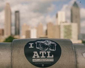 A sticker with a camera graphic and the text 'I ATL' is placed on a metal surface in the foreground. The background shows a cityscape with tall, blurred skyscrapers against a cloudy sky.