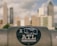 A sticker with a camera graphic and the text 'I ATL' is placed on a metal surface in the foreground. The background shows a cityscape with tall, blurred skyscrapers against a cloudy sky.