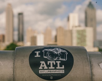 A sticker with a camera graphic and the text 'I ATL' is placed on a metal surface in the foreground. The background shows a cityscape with tall, blurred skyscrapers against a cloudy sky.