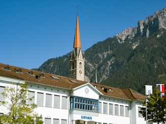 A picturesque town hall with an ornate clock tower rises against the backdrop of lush, forested mountains. The building has a red-tiled roof and white walls, accompanied by elements of modern architecture. Flags are visible beside the structure, and trees with green foliage are seen in the foreground under a clear, blue sky.