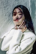 A person stands against a textured wall, wearing a cozy white sweater. Shadows of stripes are cast across their face, creating an artistic effect. The individual has long, dark hair and is touching their neck with both hands. The lighting creates a dramatic and aesthetic atmosphere.