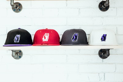 Four baseball caps are neatly lined on a wooden shelf against a white brick wall. Each cap features an embroidered bear logo. The caps are in black, red, dark gray, and white colors.