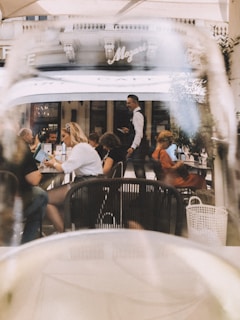 A busy outdoor café scene with people seated at tables, engaged in conversation and reading menus. The background shows a waiter in motion, serving guests. The café has a classic European facade with the name 'Mozart' prominently displayed. The image is taken through a glass, adding a soft blurring effect.
