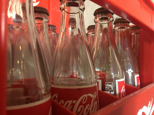 Rows of empty, clear glass bottles with red and white labels are neatly arranged in a red plastic crate. The bottles are positioned closely together, showcasing a familiar logo associated with a popular soft drink. The inner surfaces of the crate have slots that hold the bottles securely.