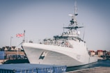 A large naval patrol ship docked at a harbor with a Union Jack flag flying at the stern. The ship is marked with the identifier P223. Nearby, there are blue cargo containers and buildings in the background under a clear sky.