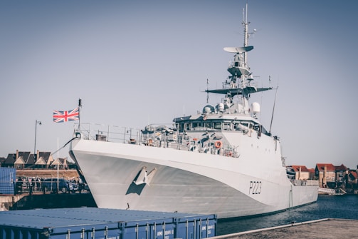 A large naval patrol ship docked at a harbor with a Union Jack flag flying at the stern. The ship is marked with the identifier P223. Nearby, there are blue cargo containers and buildings in the background under a clear sky.