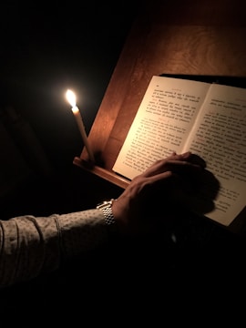 In a dimly lit setting, a candle casts a soft glow over an open book. A person's hand, wearing a patterned shirt and a watch, rests on the pages, highlighting a moment of quiet reading or study.