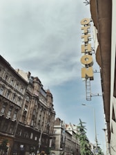 A street view featuring classic European architecture with ornate facades and a tall, narrow sign reading 'Otthon' attached to the side of a building. The sky is overcast with a hint of blue peeking through.