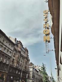 A street view featuring classic European architecture with ornate facades and a tall, narrow sign reading 'Otthon' attached to the side of a building. The sky is overcast with a hint of blue peeking through.