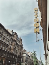 A street view featuring classic European architecture with ornate facades and a tall, narrow sign reading 'Otthon' attached to the side of a building. The sky is overcast with a hint of blue peeking through.