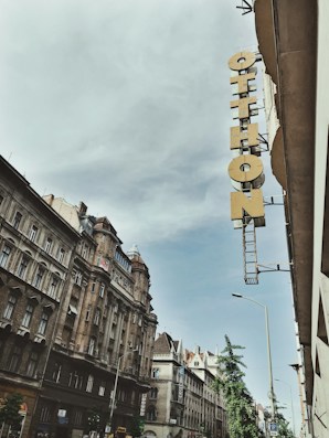A street view featuring classic European architecture with ornate facades and a tall, narrow sign reading 'Otthon' attached to the side of a building. The sky is overcast with a hint of blue peeking through.