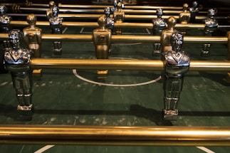 Several metallic foosball players are lined up on a green foosball table, each attached to metallic rods. The figures are painted in gold and silver with identifiable features such as crossed arms and detailed uniforms.