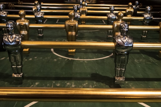 Several metallic foosball players are lined up on a green foosball table, each attached to metallic rods. The figures are painted in gold and silver with identifiable features such as crossed arms and detailed uniforms.