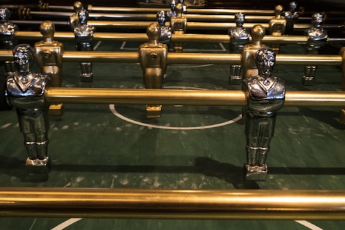 Several metallic foosball players are lined up on a green foosball table, each attached to metallic rods. The figures are painted in gold and silver with identifiable features such as crossed arms and detailed uniforms.