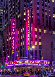 A vibrant night scene of a theater with tall neon signs displaying 'Radio City Music Hall' in bright red and blue lights. The facade is illuminated with purple and pink hues, casting a colorful glow. Below the signs, a marquee advertises upcoming events. People are walking on the sidewalk, and a yellow taxi is visible in front of the entrance. The street is bustling with activity, accentuated by several streetlights and traffic signals.