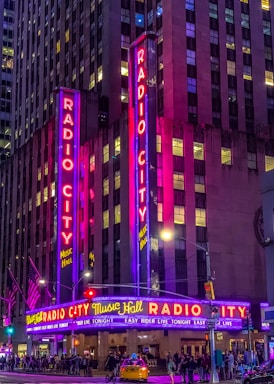 A vibrant night scene of a theater with tall neon signs displaying 'Radio City Music Hall' in bright red and blue lights. The facade is illuminated with purple and pink hues, casting a colorful glow. Below the signs, a marquee advertises upcoming events. People are walking on the sidewalk, and a yellow taxi is visible in front of the entrance. The street is bustling with activity, accentuated by several streetlights and traffic signals.