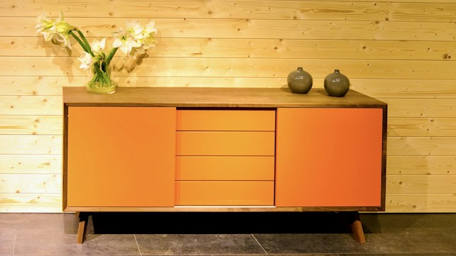 A wooden console with two orange side doors and three central drawers, placed against a light wooden panel wall. It is adorned with two dark-colored vases and a glass vase with white flowers.