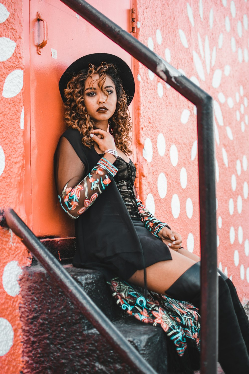 selective focus photography of of woman wearing black dress, black hat, and black thigh-high boots leaning on door