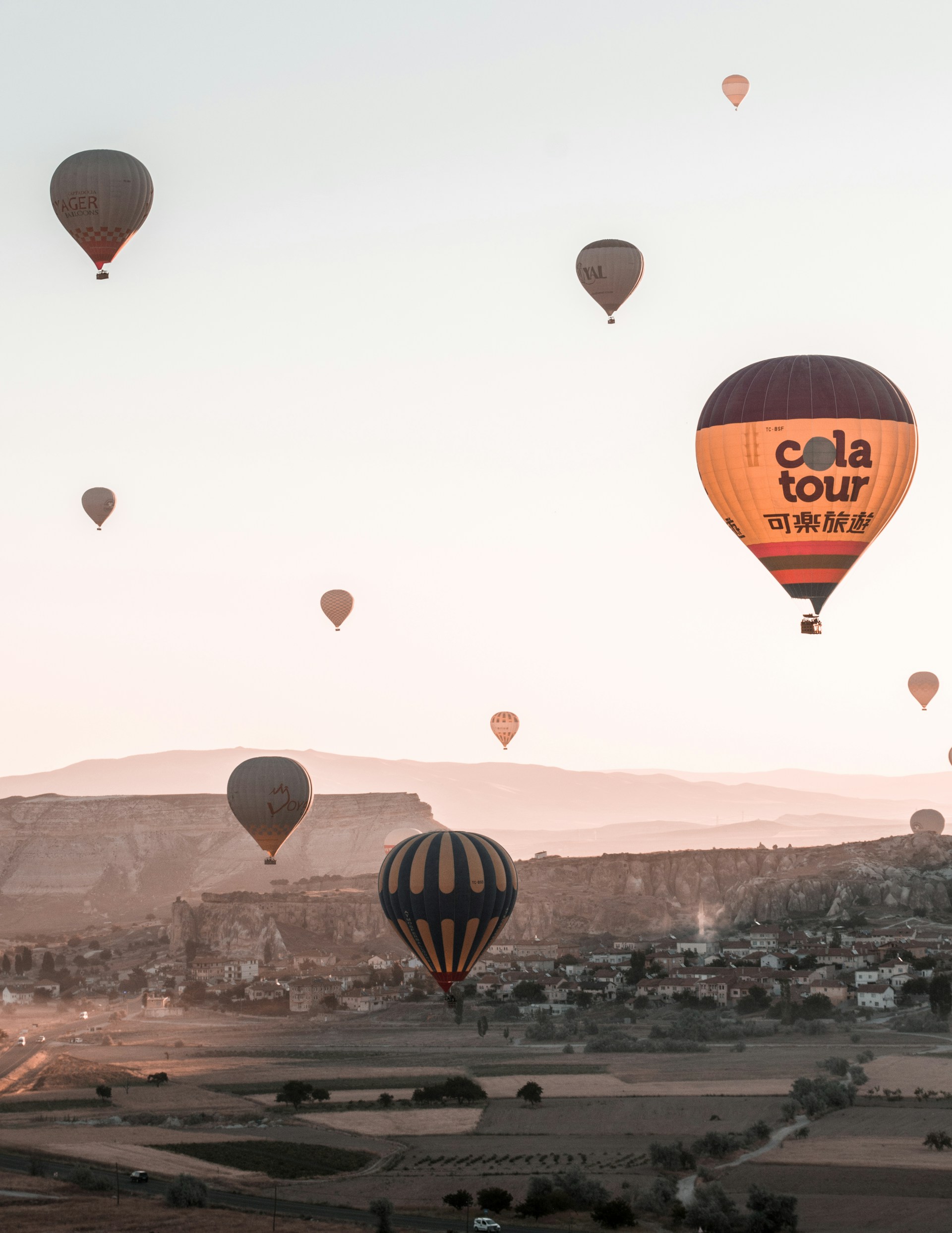 Hot air balloons are floating across a landscape during either sunrise or sunset, with gentle hills and scattered greenery below. The sky is a soft pastel, and the light casts a warm glow over the scene, highlighting the rocky terrain and small settlements below.