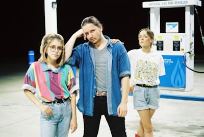 Three individuals standing in front of a natural gas pump at a station. The person on the left wears glasses and a colorful striped shirt, while the central figure dons a denim shirt over a gray t-shirt. The person on the right is dressed casually in a graphic t-shirt and denim shorts. The scene is set at night.