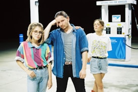 Three individuals standing in front of a natural gas pump at a station. The person on the left wears glasses and a colorful striped shirt, while the central figure dons a denim shirt over a gray t-shirt. The person on the right is dressed casually in a graphic t-shirt and denim shorts. The scene is set at night.
