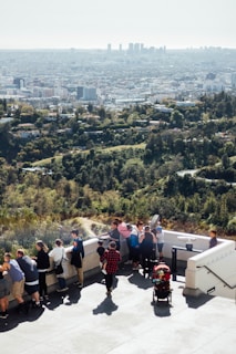 A group of people are gathered on a viewing platform overlooking a sprawling city skyline in the distance. The foreground shows a variety of people, including adults and children, leaning against the railing and looking out at the view. A child in a red stroller is also visible, and people appear to be enjoying the sunny day. The background is dominated by the green landscape transitioning into the cityscape.