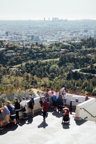 A group of people are gathered on a viewing platform overlooking a sprawling city skyline in the distance. The foreground shows a variety of people, including adults and children, leaning against the railing and looking out at the view. A child in a red stroller is also visible, and people appear to be enjoying the sunny day. The background is dominated by the green landscape transitioning into the cityscape.